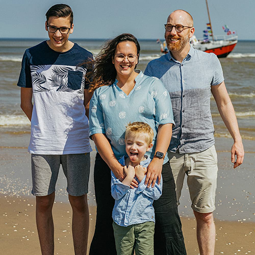 Picture of a family of 4 posing on the beach with the sea behind them. A mother stands in the middle with her teenage son on the left and husband on the right. In front of her stands her toddler sticking out his tongue while she gently has her arms resting on his shoulders.